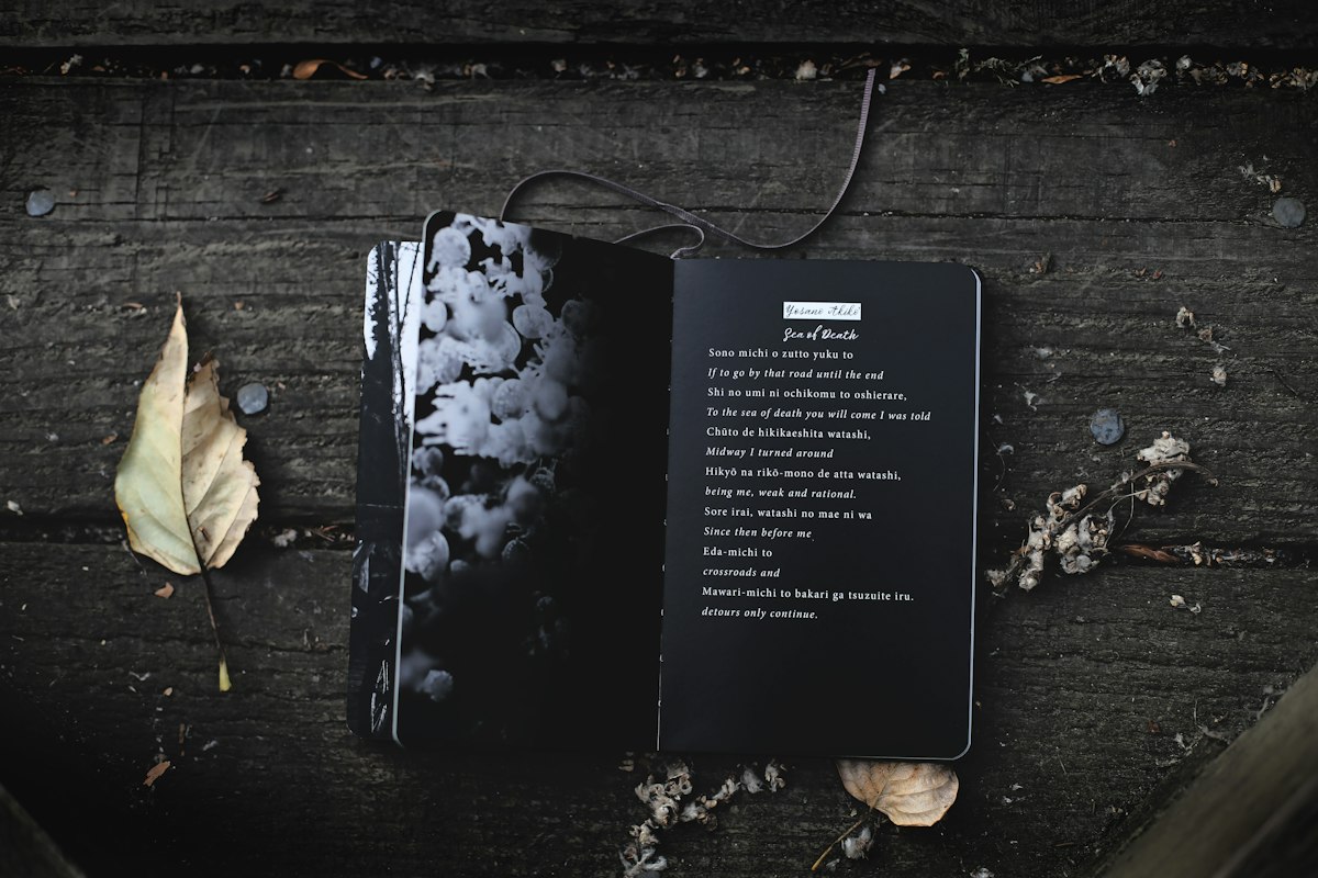 An open black notebook with a poem printed in white text, lying on weathered wooden boards among dry leaves