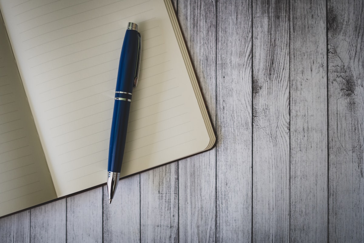 A blue click pen resting on a brown wooden table beside a notebook