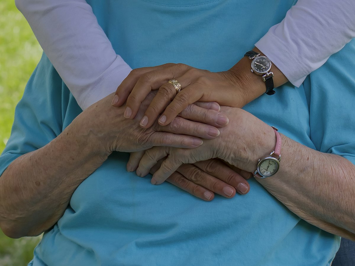 A younger person's arms wrapped around an older person from behind, their hands clasped together