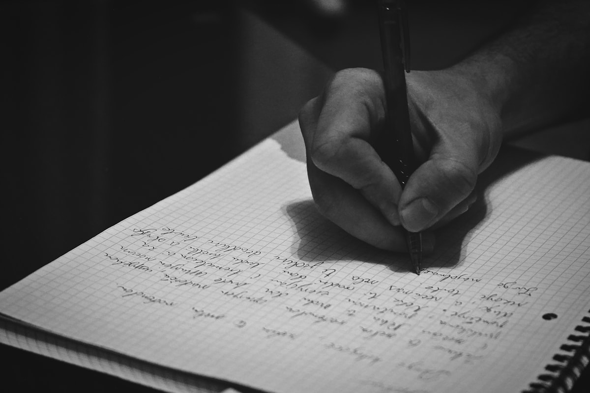 An open notebook on a wooden desk beside a dim lamp, a pen resting on an unfinished page