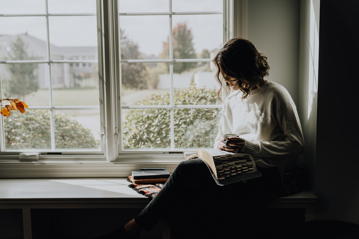 A woman sitting quietly by a window with a book, embodying the reflective solitude of introvert life