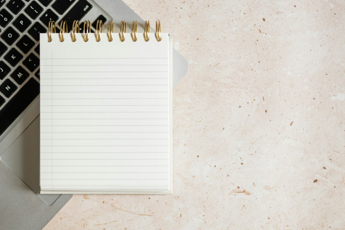 A notepad resting on top of a laptop beside a keyboard, suggesting a writer's working desk