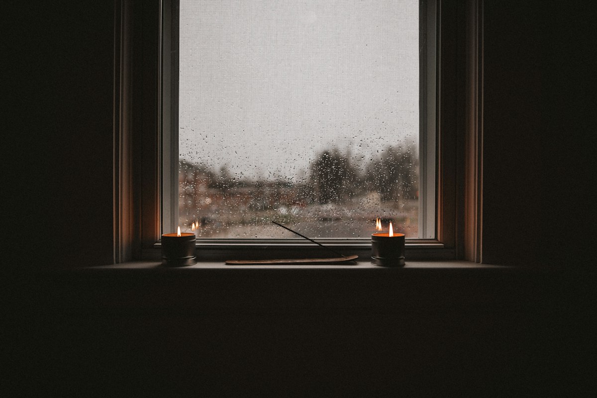 A rain-streaked window at dusk with two small candles lit on the sill