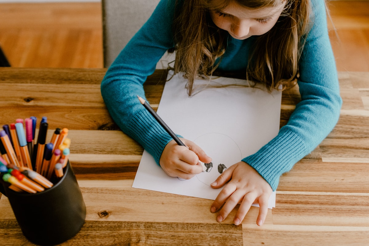 A child's hand drawing in a notebook at a kitchen table beside coloured pencils