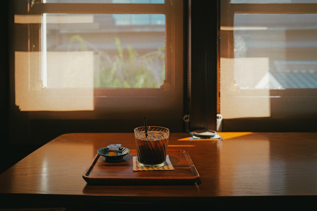 A cold drink on a wooden table beside a window in soft daylight, a quiet moment to sit down and write again