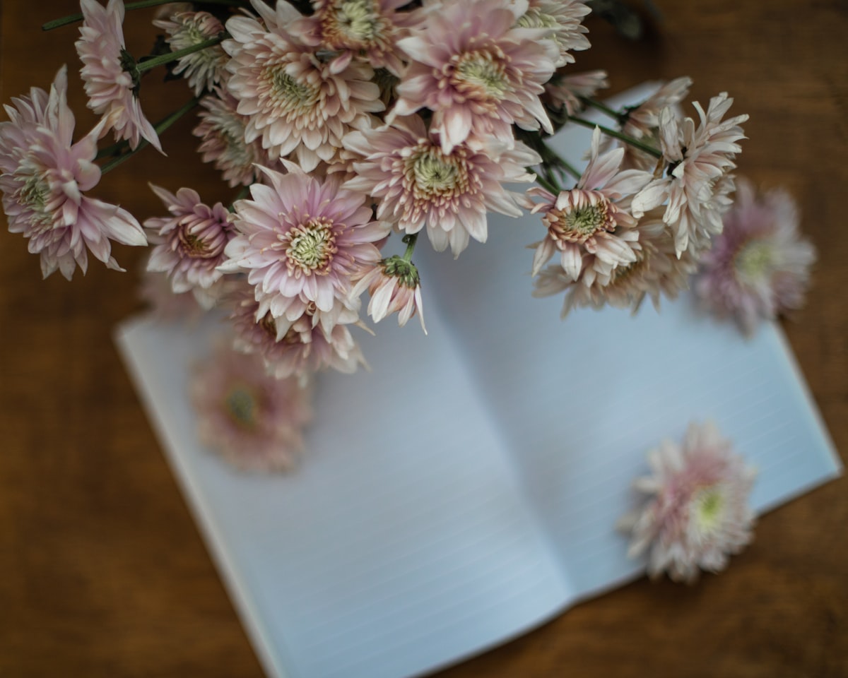 An open notebook on a wooden table beside a vase of fresh spring flowers and an open window letting in soft daylight
