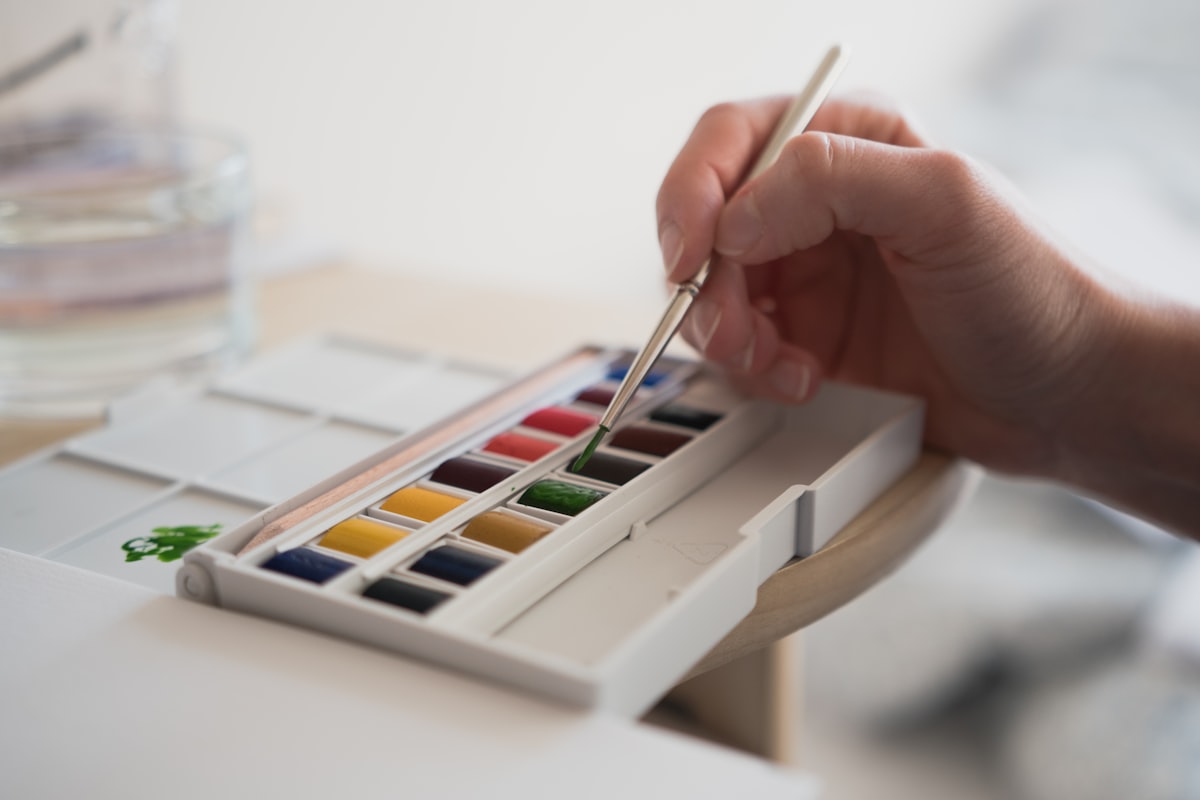 A hand loading a fine brush with green paint from an open watercolor pan set beside a jar of water